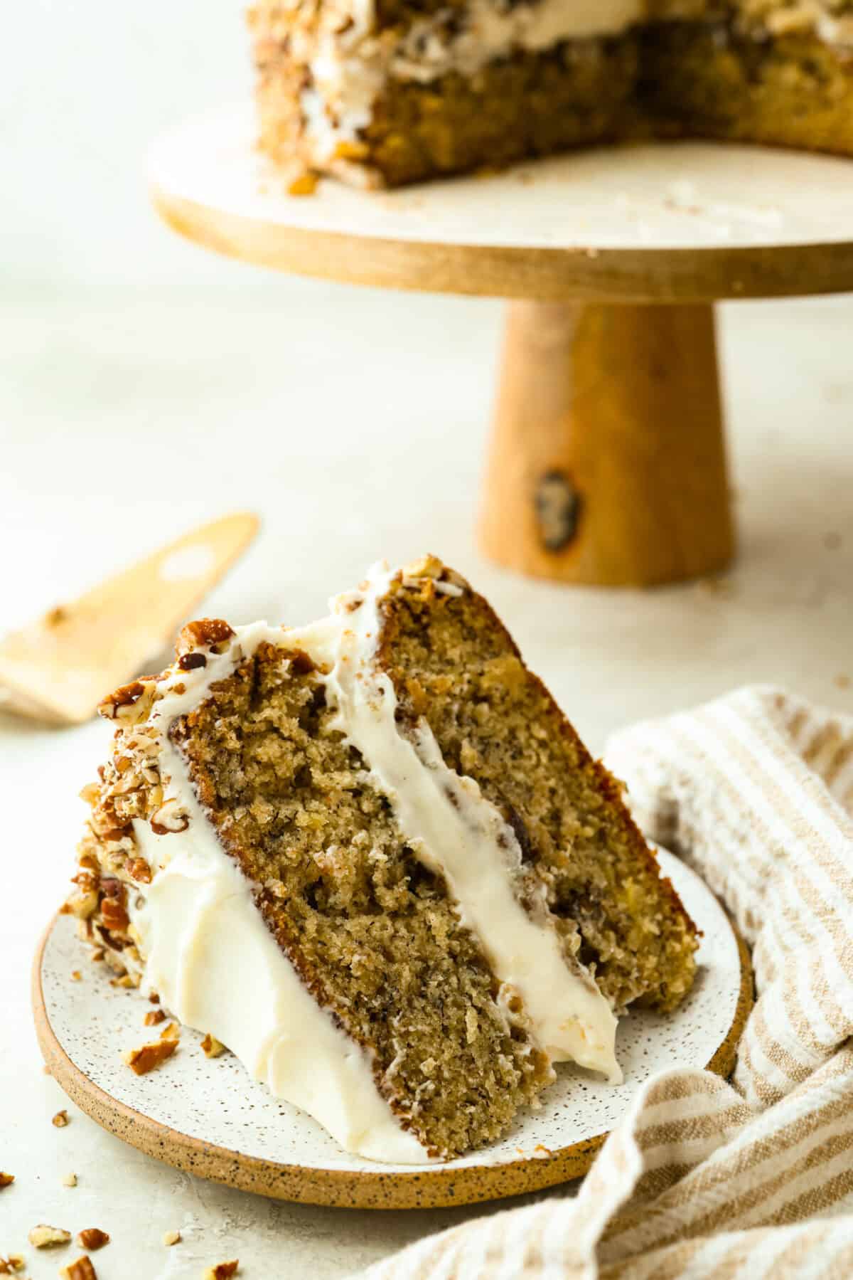 If you’ve never had hummingbird cake, just know it’s one of those cakes people take a bite of and immediately ask for the recipe. It’s soft, sweet, packed with banana and pineapple, and topped with a cream cheese frosting that is to die for! Slice of hummingbird cake on a plate next to the cake stand with the rest of the cake.