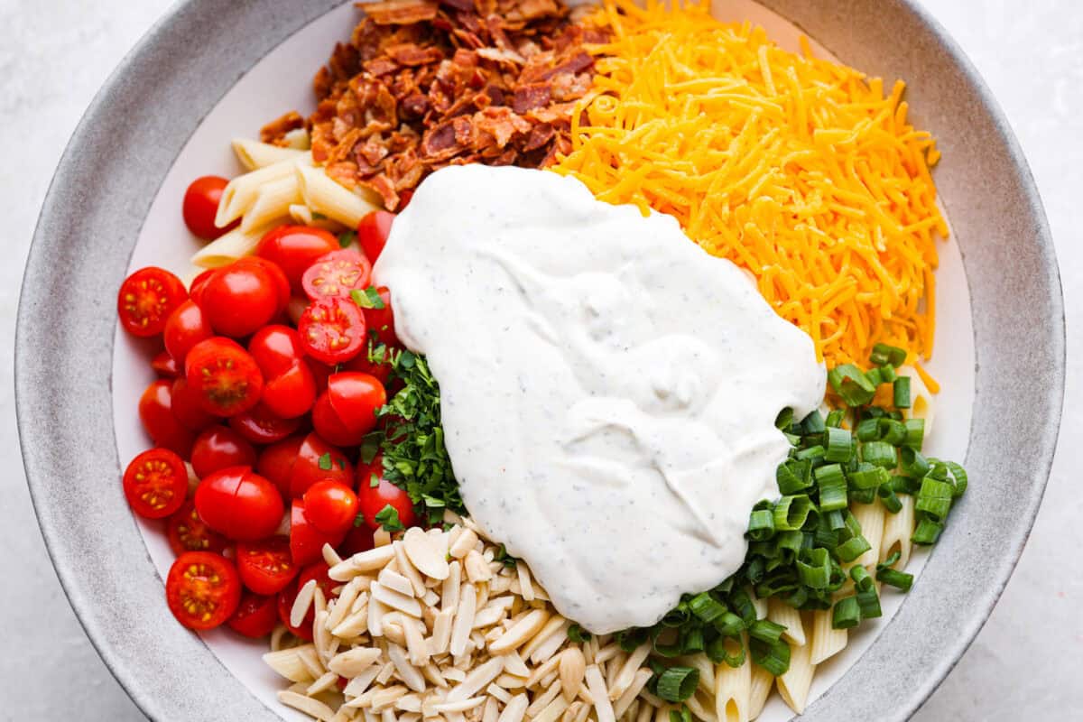 If there’s one side dish that disappears fast, it’s this million dollar pasta salad. It’s packed with crispy bacon, sharp cheddar, fresh veggies, and a creamy dressing that ties it all together. It’s sure to steal the spotlight at any table! Dressing added to the top of the salad ingredients ready to be mixed in.