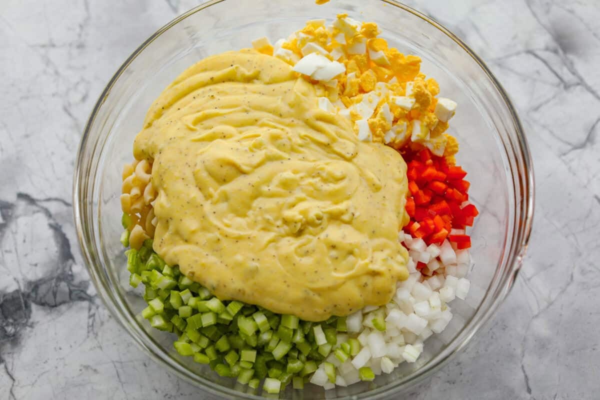 There’s something delightfully old school about Amish pasta salad. The sweet, tangy dressing, the macaroni, the chopped eggs, the crunchy celery. It tastes like the kind of recipe that’s been living in a handwritten church cookbook for decades. Dressing added to the bowl with pasta salad ingredients.