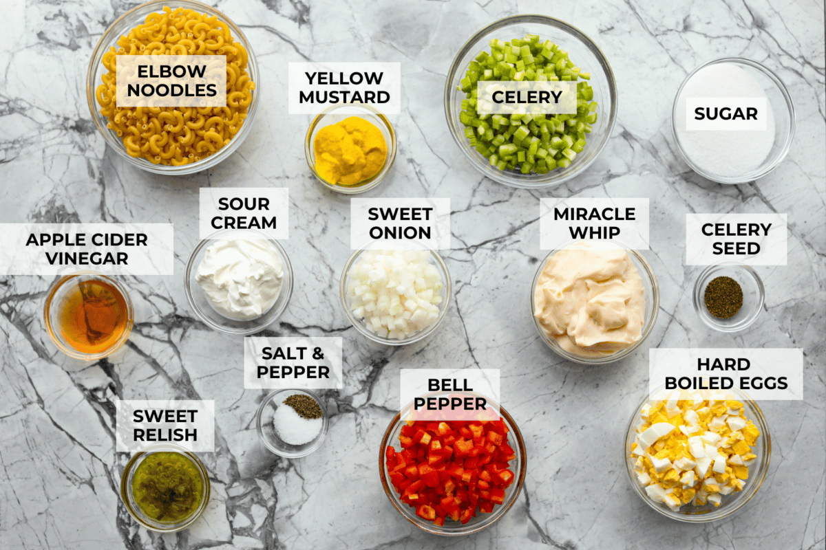 There’s something delightfully old school about Amish pasta salad. The sweet, tangy dressing, the macaroni, the chopped eggs, the crunchy celery. It tastes like the kind of recipe that’s been living in a handwritten church cookbook for decades. Overhead shot of labeled amish macaroni salad ingredients.
