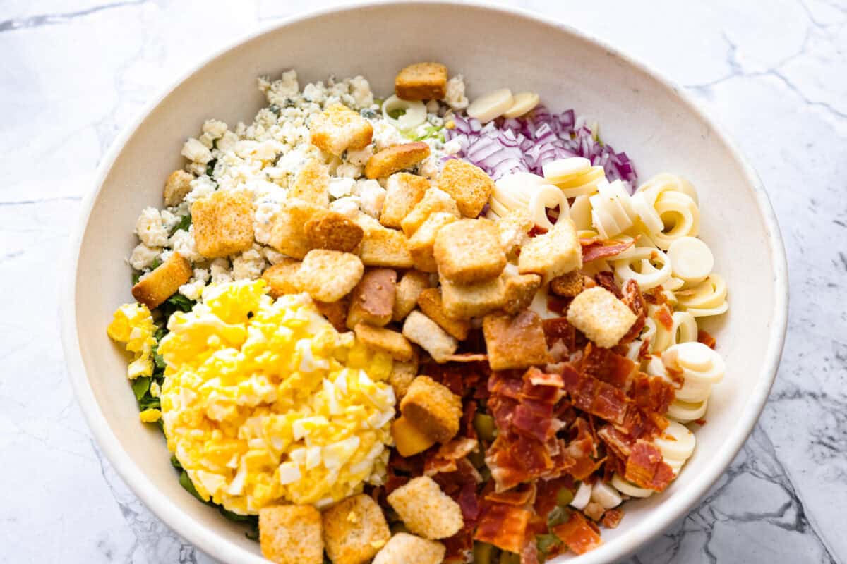 Ruth's Chris chopped salad is a steakhouse legend, and now you can make it at home for a fraction of the price. Crisp greens, smoky bacon, bold blue cheese, and a tangy lemon basil dressing come together in one showstopping bowl that tastes just like the real thing. Bowl with all of the different ingredients for the salad.