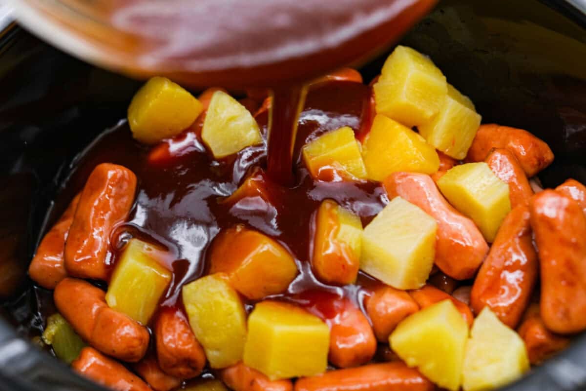 Don't be surprised if these Hawaiian Lil' Smokies steal the show! Slow cooked in a sweet and tangy pineapple BBQ sauce, they're bite-sized, irresistible, and dangerously easy to snack on. Sauce being poured over the pineapple and lit'l smokies in the slow cooker.