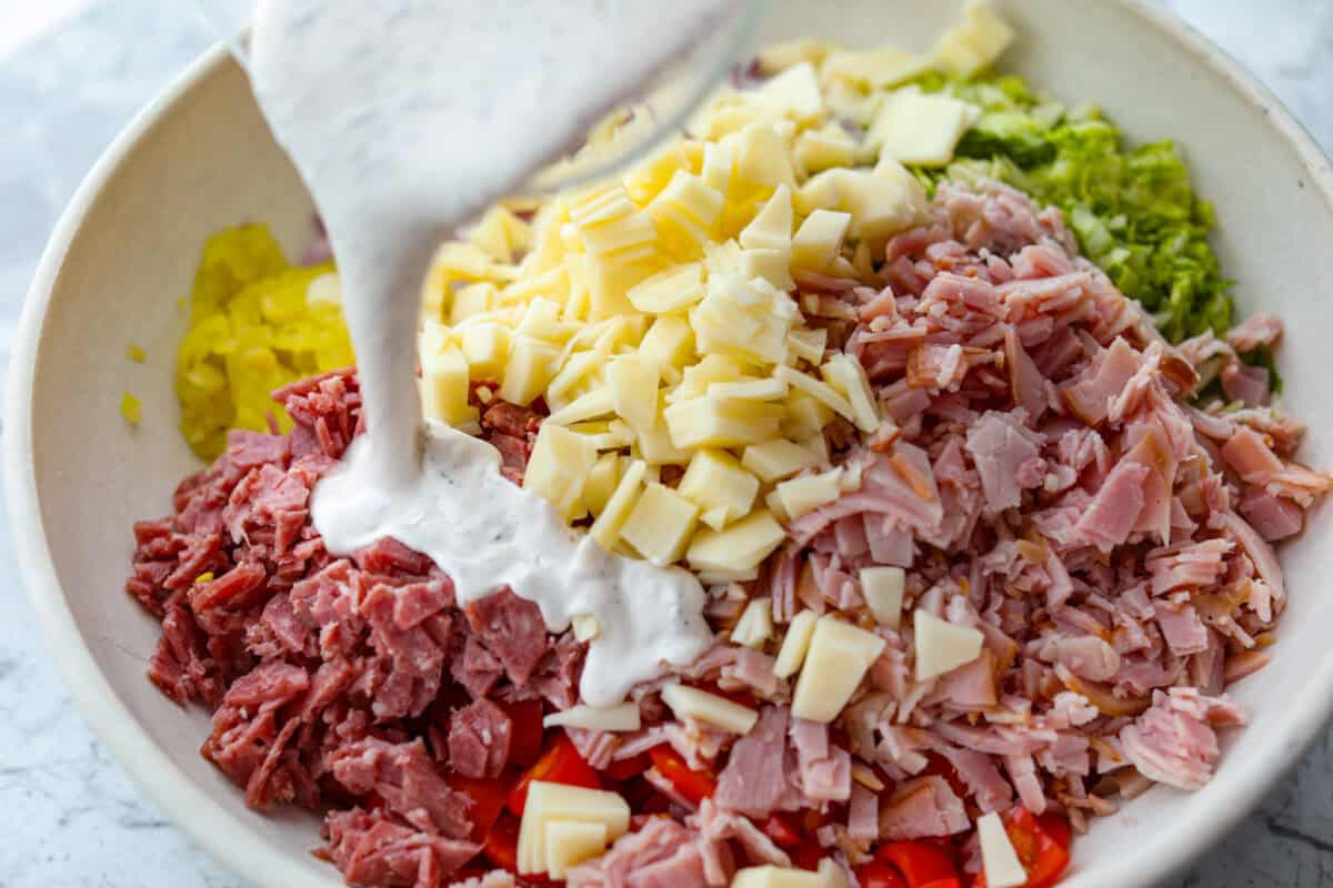 If you're a fan of deli sandwiches, you will love this sub in a tub dip! Made with deli meats, cheese, crisp veggies, and a creamy tangy dressing, it tastes just like your favorite sandwich shop sub in dip form! Dressing being poured over the sub in a tub dip ingredients.
