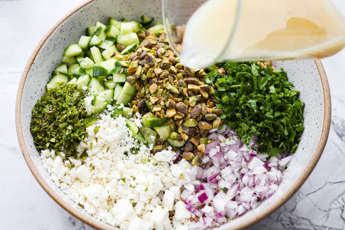 This Jennifer Aniston Salad is light, fresh, and perfect for summer lunches. Itโs packed with veggies, chickpeas, herbs, pistachios, and feta, then tossed in a simple lemon dressing that makes everything taste bright and delicious. Someone pouring the dressing over the salad ingredients.