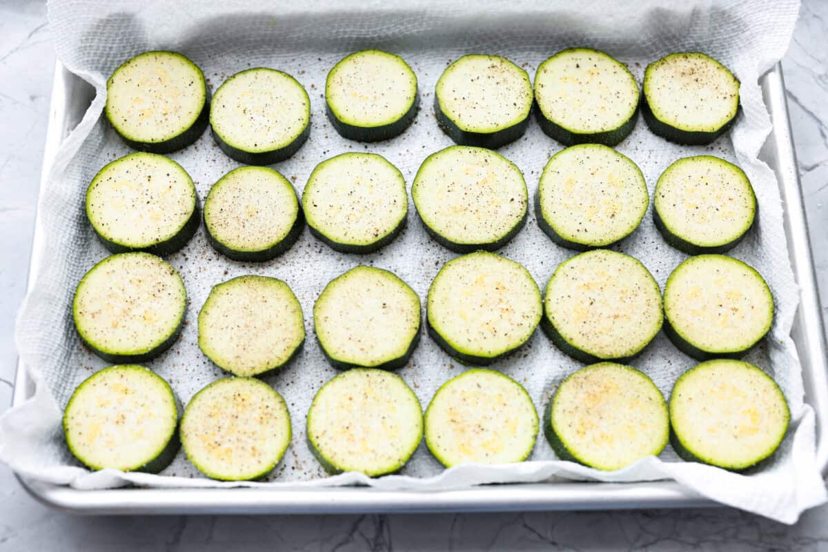 When my garden is overflowing with zucchini, this crispy Parmesan fried zucchini is one of my favorite things to make. It’s ready in just 15 minutes and is perfect as a quick appetizer, snack, or easy side with ranch, marinara, or fry sauce. Zucchini slices on parchment paper on a baking sheet with salt and pepper sprinkled over them