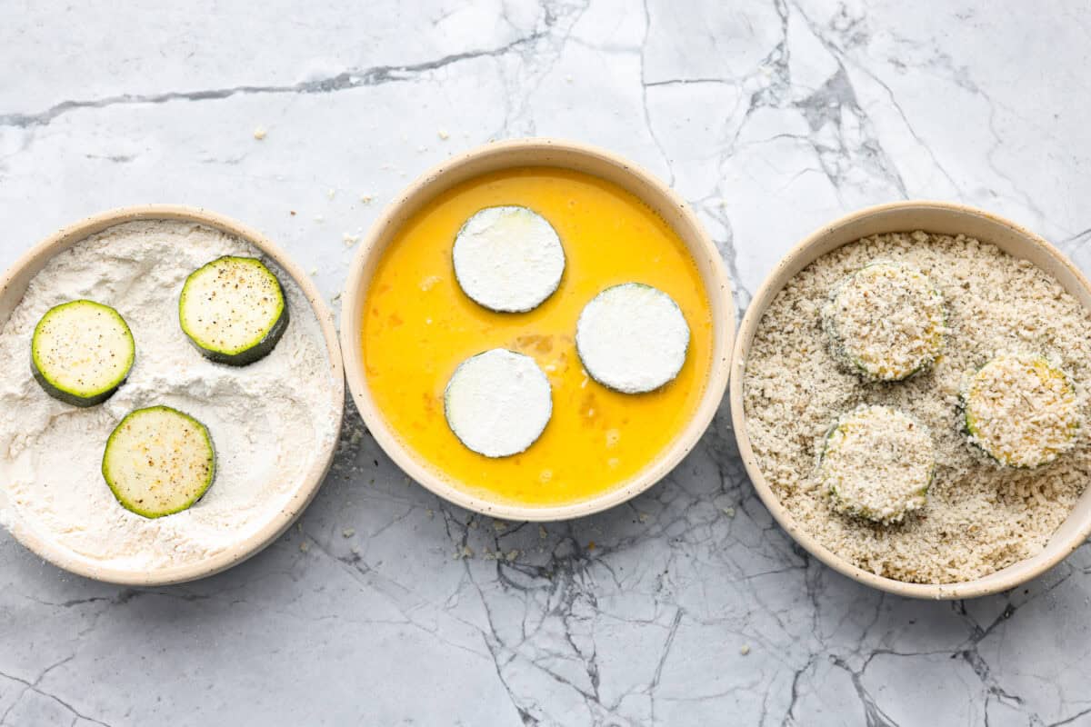 When my garden is overflowing with zucchini, this crispy Parmesan fried zucchini is one of my favorite things to make. It’s ready in just 15 minutes and is perfect as a quick appetizer, snack, or easy side with ranch, marinara, or fry sauce. Zucchini slices in each of the dredging bowls.