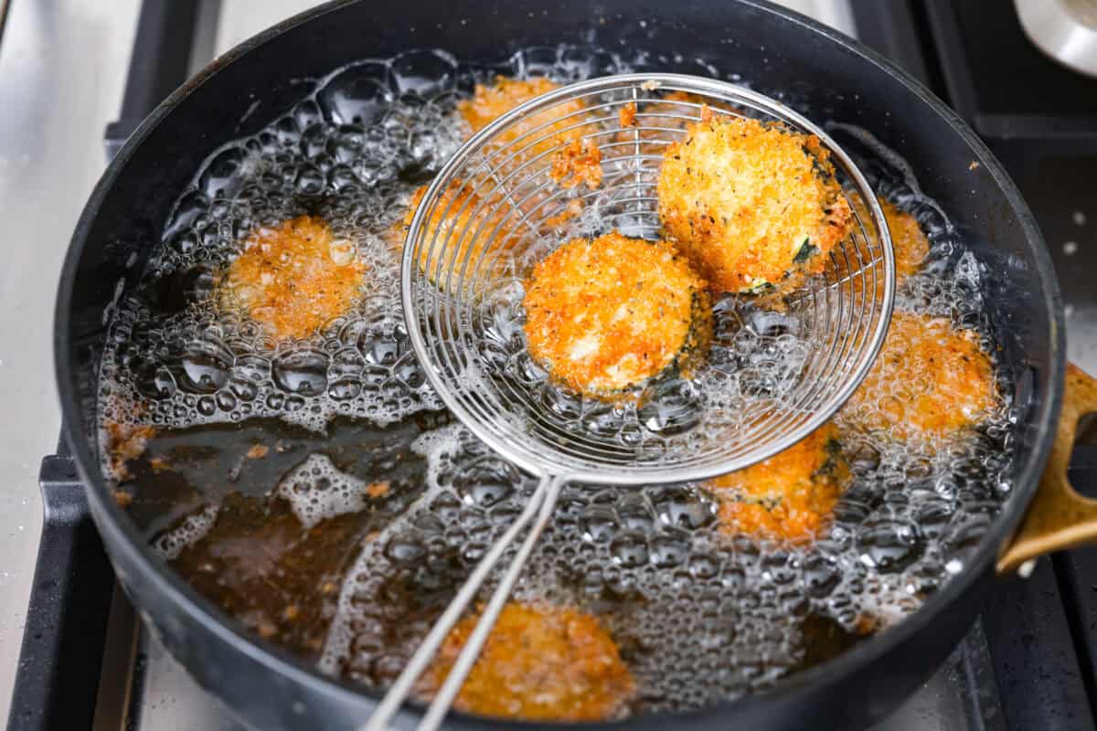 Someone lifting the fried zucchini out of the fryer with a spider strainer.