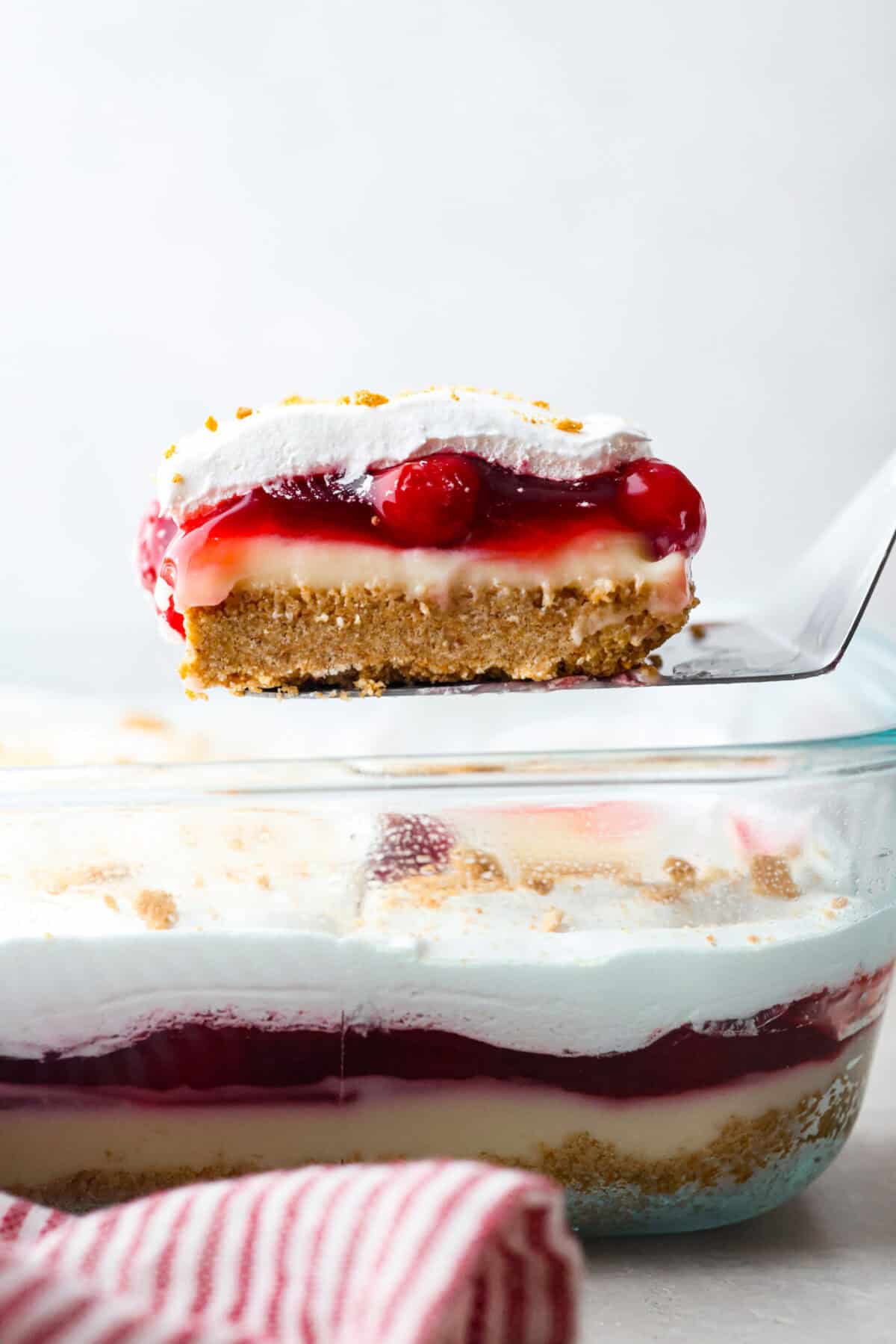 My cherry cheesecake lush is rich, creamy, and incredibly easy to make. With simple layers of graham cracker crust, cheesecake filling, juicy cherries, and whipped topping, it's the perfect no-bake dessert for any occasion. Someone lifting a slice of cherry cheesecake lush out of the glass dish.