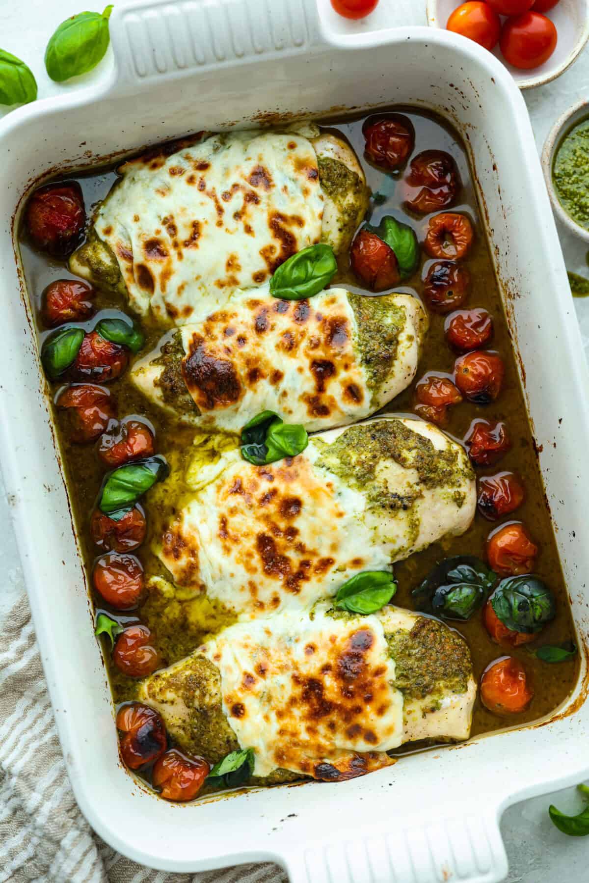 This pesto chicken is the kind of dinner you can assemble right in the baking dish and let the oven take it from there. Add the chicken, pesto, mozzarella, and tomatoes to one pan, bake until tender and bubbly, and dinner is done. Baking dish with baked pesto chicken surrounded by cherry tomatoes.