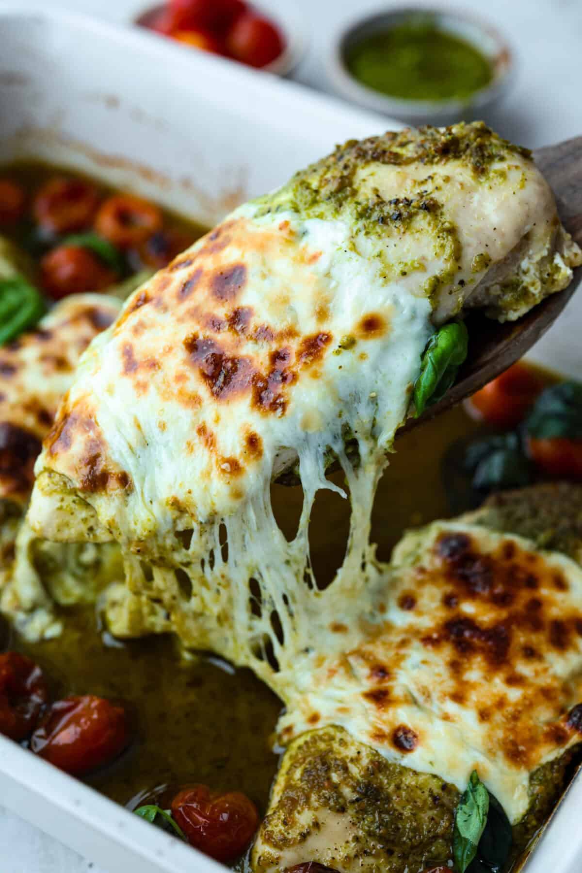 This pesto chicken is the kind of dinner you can assemble right in the baking dish and let the oven take it from there. Add the chicken, pesto, mozzarella, and tomatoes to one pan, bake until tender and bubbly, and dinner is done. Someone lifting out a cheesy baked pesto chicken breast from the baking dish.
