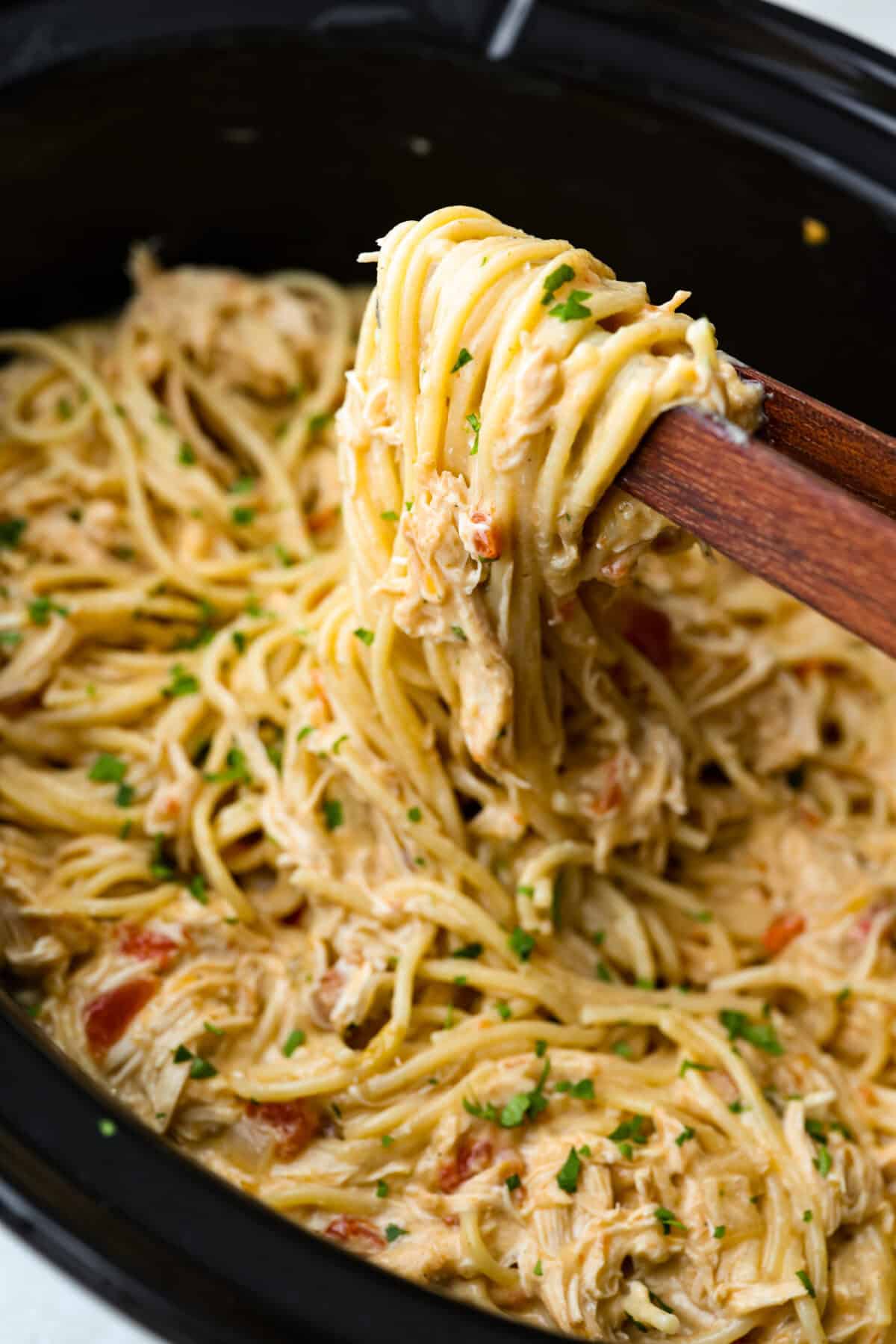 Someone lifting up a scoop of crockpot chicken spaghetti with wooden tongs. 