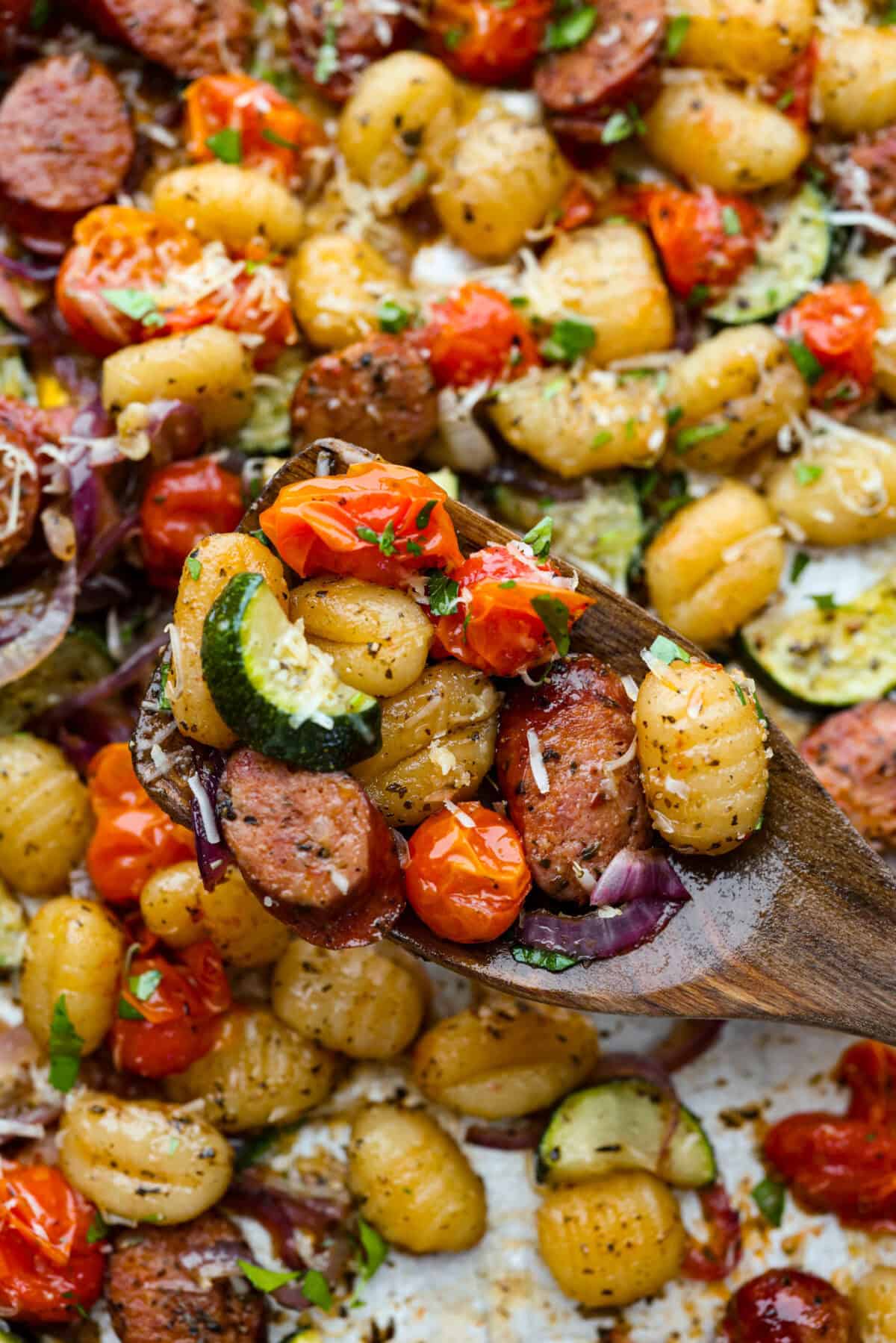 This sheet pan gnocchi is about as easy as dinner gets! Everything goes onto one pan with olive oil and Italian seasoning, then roasts until the gnocchi is crispy and the veggies are tender. Finish it with Parmesan and dig in! Close up of a scoop of sheet pan gnocchi on a wooden spoon.